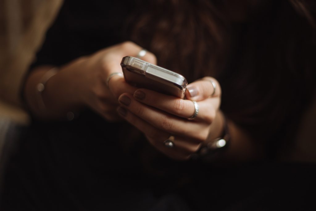 A lady holds her mobile phone. We see her hands upclose with rings on her fingers. The background is dark.